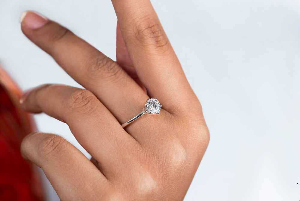 Model hand displays a sparkling 1 carat moissanite stone solitaire ring with a silver band on a finger.