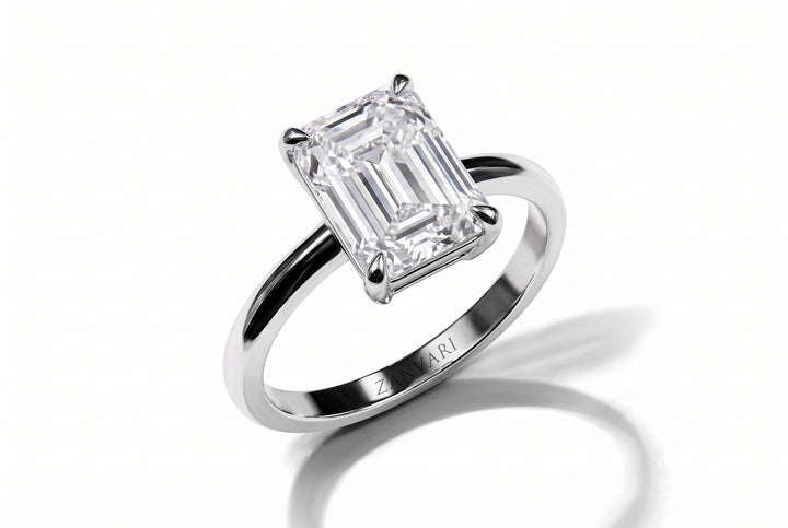 An emerald-cut diamond solitaire ring in a polished silver-colored metal band, shown on a clean white background with a soft shadow.