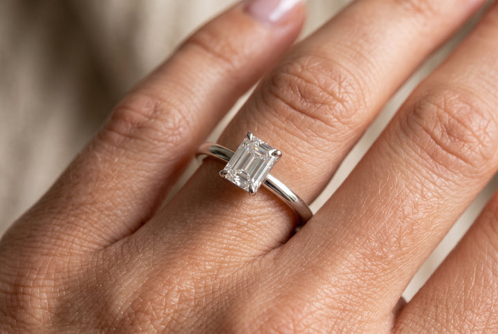 Macro close-up of a hand wearing a delicate white gold solitaire engagement ring featuring a dainty emerald-cut lab diamond in a four-prong setting.