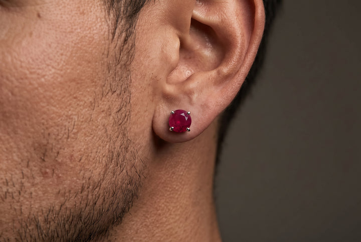Close-up portrait view of a man’s earlobe wearing a round-cut, deep red opaque ruby stud earring set in white gold prongs.   
