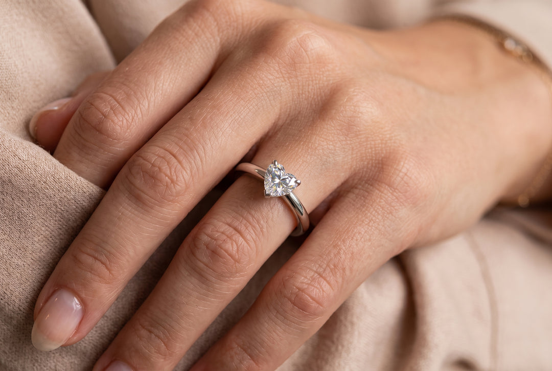 A detailed, close-up photograph focuses on a person's left hand, viewed from an upper three-quarter angle, resting against a soft, textured beige fabric. A slender white-gold or platinum ring, adorned with a prominent heart-shaped diamond, is worn on the ring finger. The heart-shaped diamond is set with its point facing down the finger, catching the light in a multi-faceted brilliant sparkle. The band is smooth and polished. The fingernails are neatly trimmed and natural, with the cuticle area and a partial