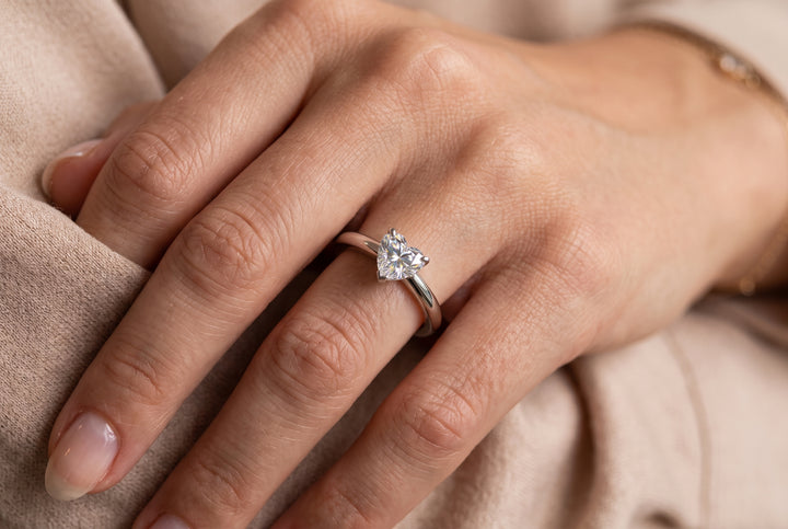 A detailed, close-up photograph focuses on a person's left hand, viewed from an upper three-quarter angle, resting against a soft, textured beige fabric. A slender white-gold or platinum ring, adorned with a prominent heart-shaped diamond, is worn on the ring finger. The heart-shaped diamond is set with its point facing down the finger, catching the light in a multi-faceted brilliant sparkle. The band is smooth and polished. The fingernails are neatly trimmed and natural, with the cuticle area and a partial