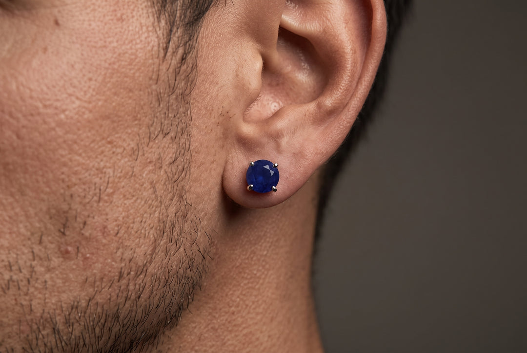 A close-up of a man's ear wearing a round, deep blue sapphire gemstone stud earring in a prong setting against a dark gray background.
