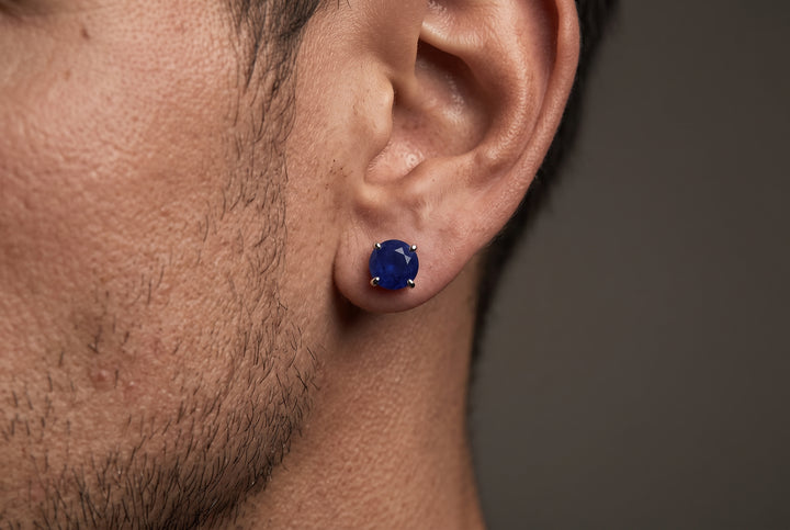 A close-up of a man's ear wearing a round, deep blue sapphire gemstone stud earring in a prong setting against a dark gray background.