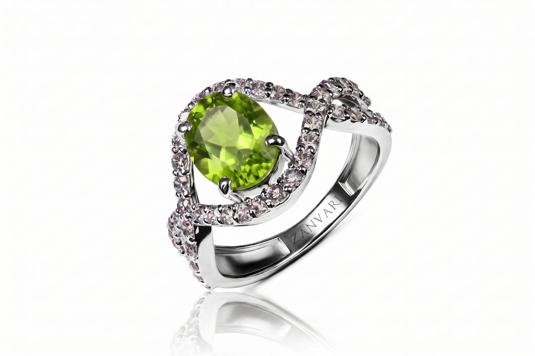 A silver ring featuring a large oval green peridot gemstone surrounded by a halo of small clear stones, with a twisted band accented by pavé-set clear stones. The brand name 'ZANVARI' is engraved on the inner band.