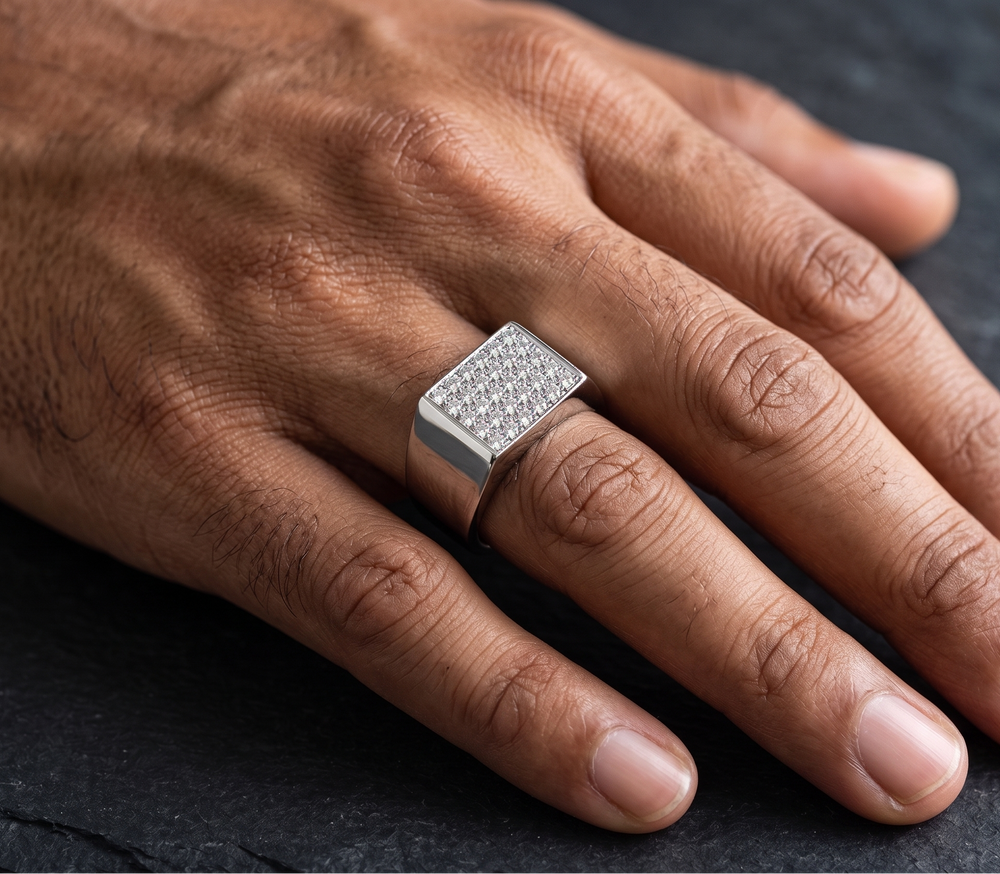 A close-up of a hand wearing a wide silver signet ring with a square face paved with small clear stones, resting on a dark slate surface.