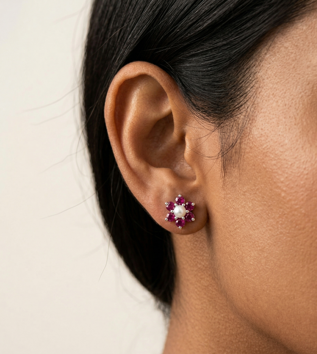 Close-up of an ear wearing a floral earring with red  ruby and pearl stones on a neutral background