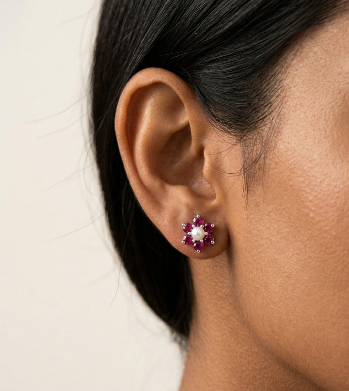 Close-up of an ear wearing a floral earring with red  ruby and pearl stones on a neutral background