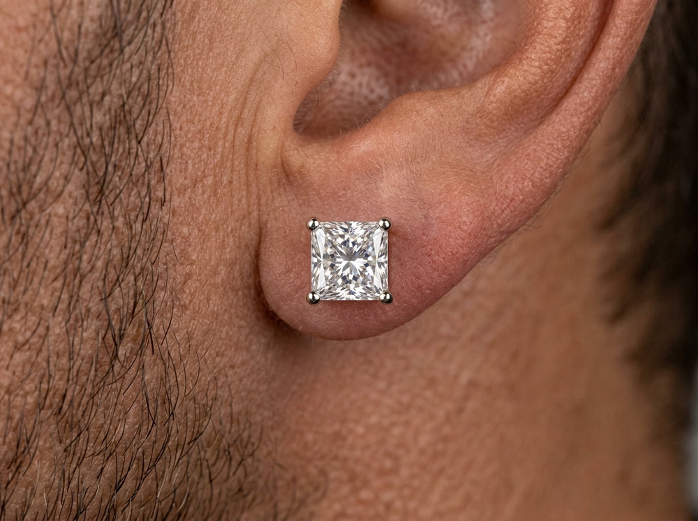 Extreme macro photograph showing a square princess-cut diamond stud earring with four white metal prongs worn centered on a man's earlobe.