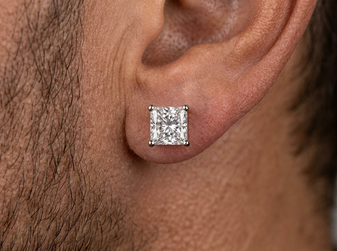 Extreme macro photograph showing a square princess-cut diamond stud earring with four white metal prongs worn centered on a man's earlobe.