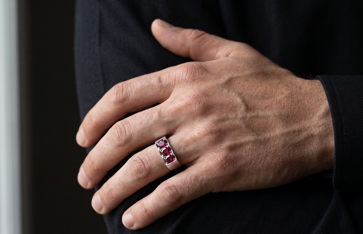 A close-up of a man's hand wearing a silver band ring set with three marquise-cut red ruby gemstones, set against his dark clothing.