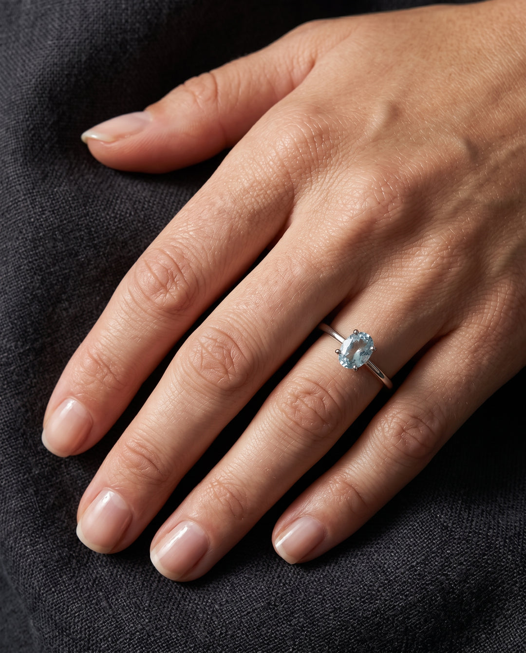 A close-up of a hand wearing a silver ring with a round light blue aquamarine gemstone, resting on a dark textured fabric background.