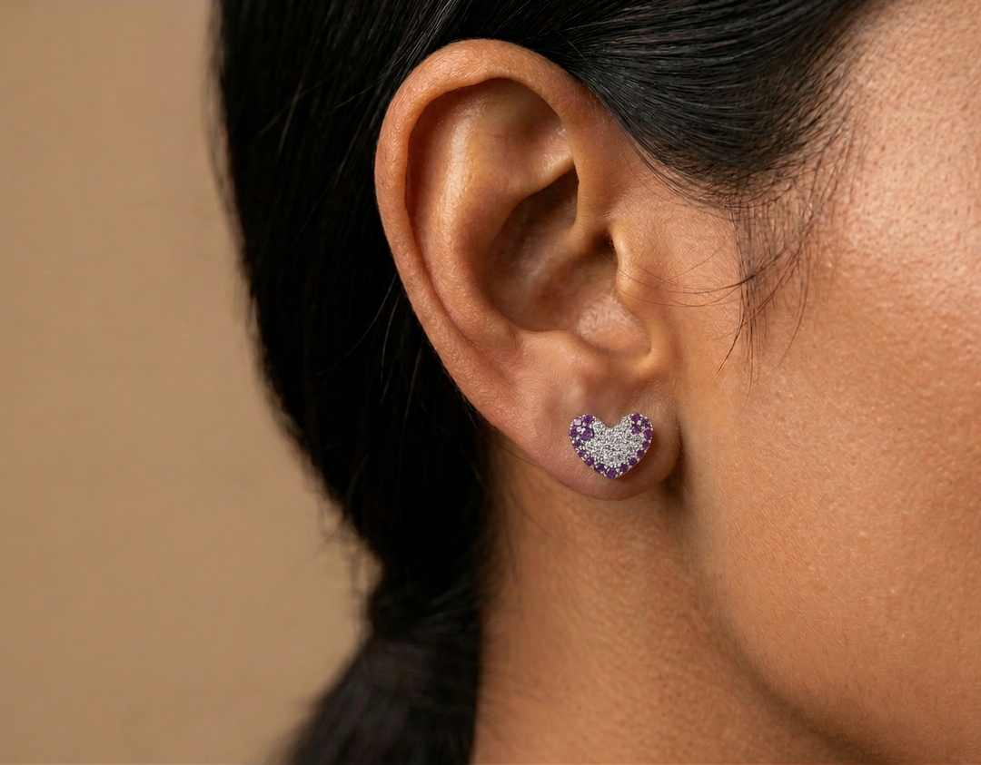 Close-up of an ear wearing a heart-shaped earring with ruby stones against a neutral background