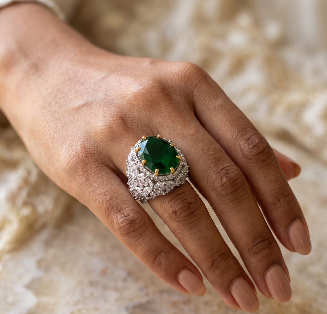 A close-up of a hand wearing an elegant ring with a large oval-cut green jade center stone and a silver pavé-set band, set against a soft beige stone background.