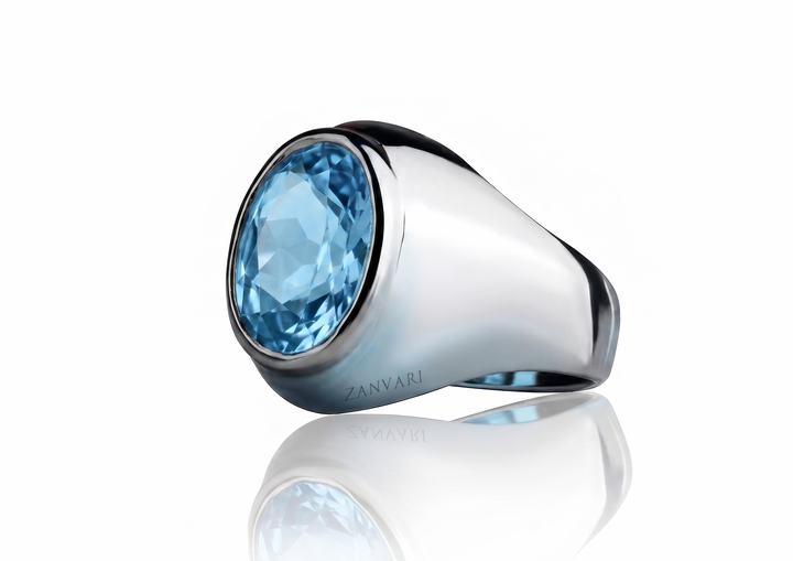 Silver masculine signet ring with an oval-cut light blue Swiss topaz stone set in a sleek bezel, displayed on a reflective white studio background with the logo "ZANVARI".