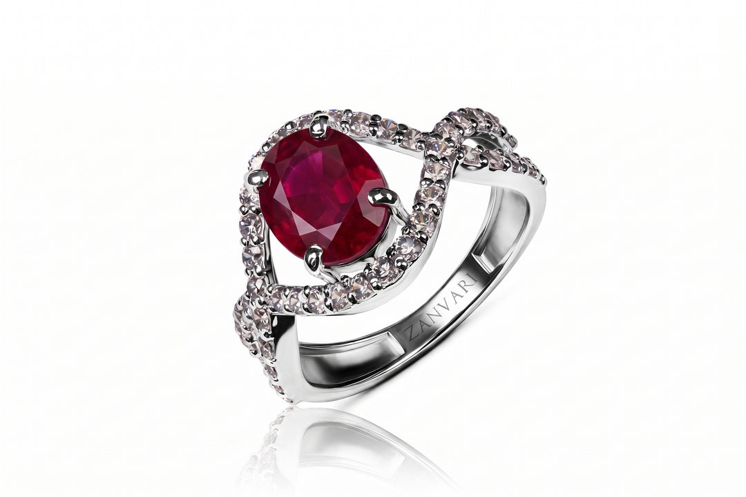 A silver-toned ring featuring a large oval deep red ruby center stone, surrounded by a halo of small clear pavé stones and a twisted band with an engraved NVAR logo, shown on a white background with a soft reflection.