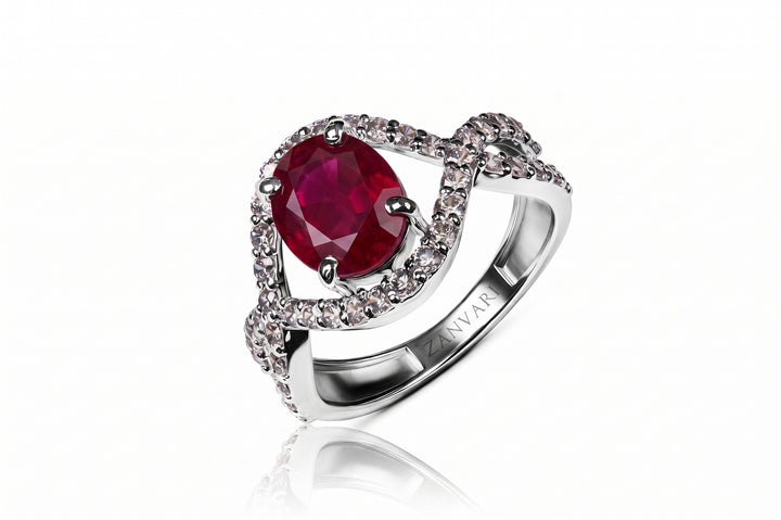 A silver-toned ring featuring a large oval deep red ruby center stone, surrounded by a halo of small clear pavé stones and a twisted band with an engraved NVAR logo, shown on a white background with a soft reflection.