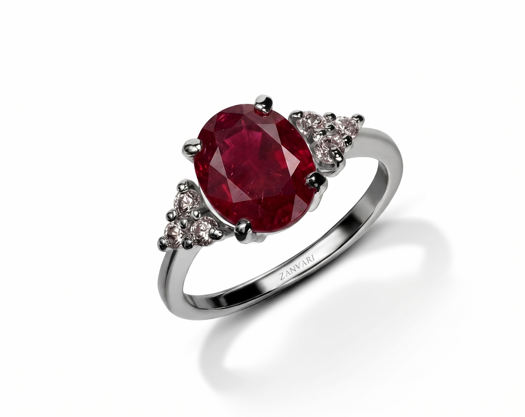 A luxury silver ring featuring a large oval-cut deep red ruby center stone, accented by small round moissanite in triangular clusters on either side of the band. The polished silver band is inscribed with the brand name 'ZANVARI'.