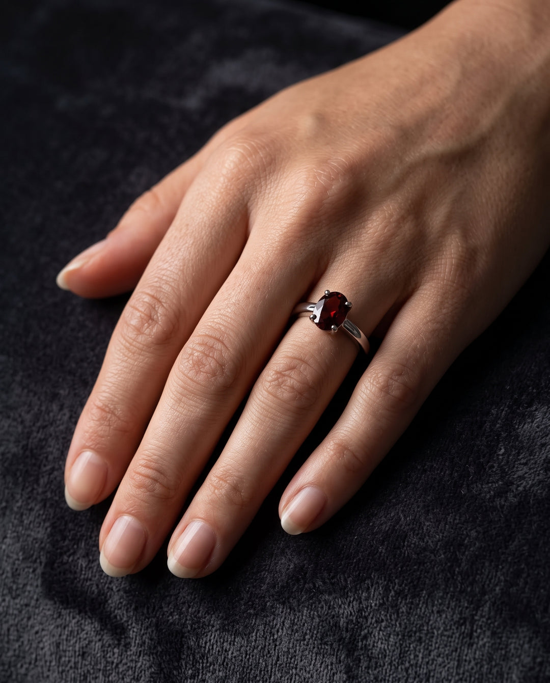 A high-fashion editorial close-up of an elegant woman’s hand with neatly manicured natural nails resting diagonally across dark charcoal drapery. A polished silver solitaire ring set with a faceted deep red oval garnet is worn on the ring finger, highlighted by soft, directional window light with natural shadows and authentic skin texture.