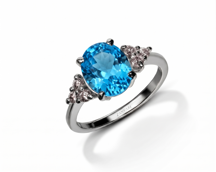 A silver ring featuring a large oval-cut bright blue swiss topaz gemstone in the center, accented by two small triangular clusters of clear stones on either side of the band. The brand name 'Zanvari' is engraved on the inner band.