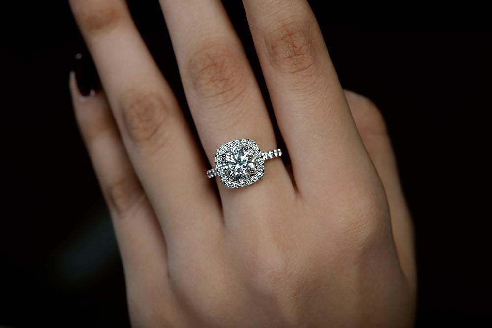 A hand elegantly displays a silver ring with a square halo of small diamonds around a central Moissanite stone