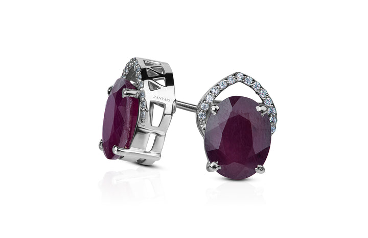 A pair of white gold-plated sterling silver stud earrings featuring a large natural ruby center stone with smaller cubic zirconia side stones.