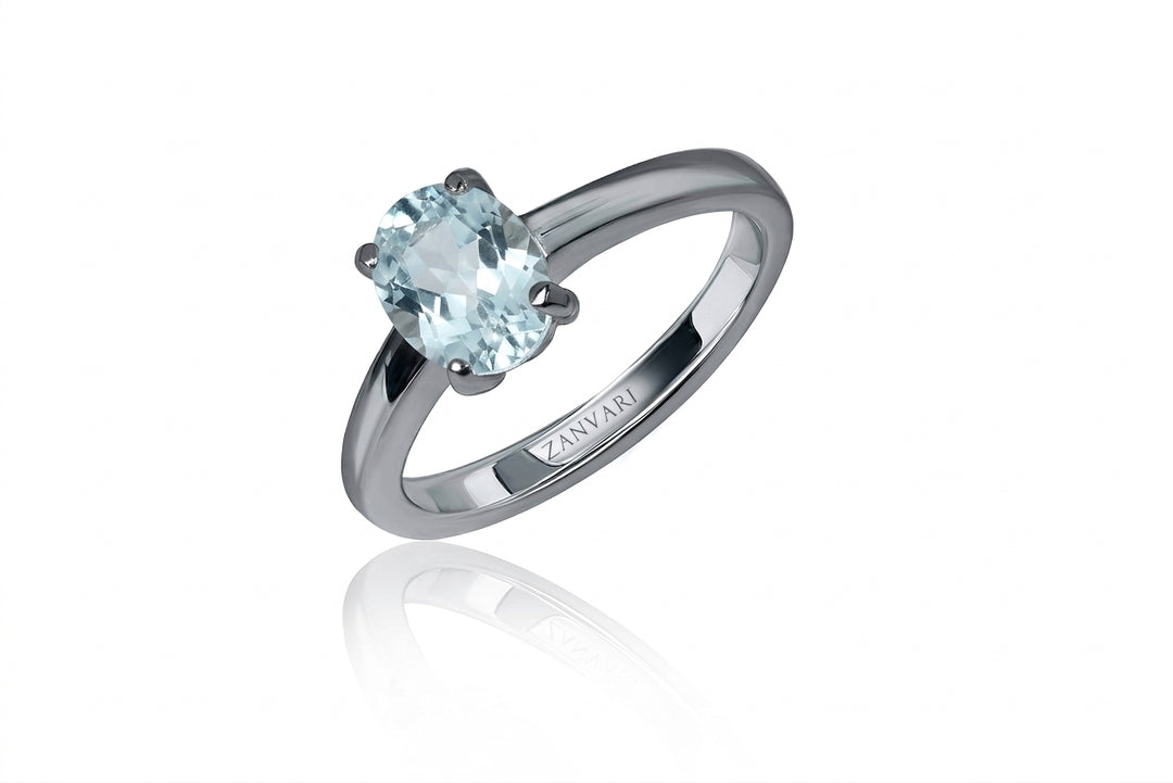 A studio product shot of a silver solitaire ring featuring a faceted oval icy-blue aquamarine held by four prongs, angled upright with a soft mirror reflection on a pure white surface.