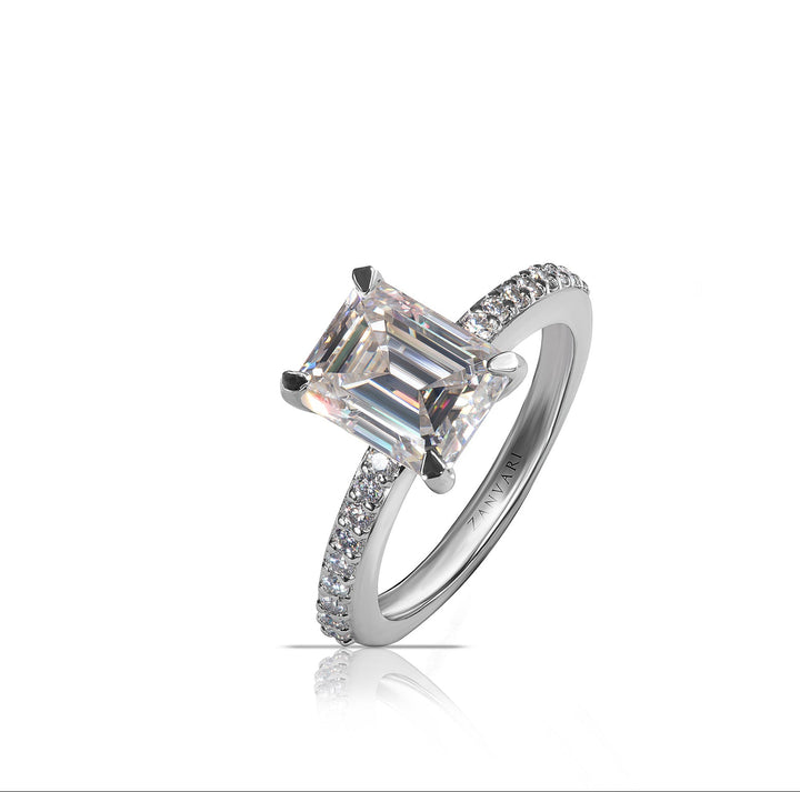 An emerald-cut diamond engagement ring in a silver-colored metal band with pavé-set accent diamonds, shown on a clean white background with a soft reflection below.