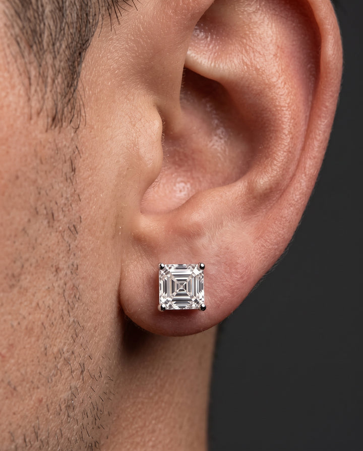 A close-up of a man's ear wearing a single square brilliant-cut diamond stud earring in a silver-toned setting against a dark gray background.