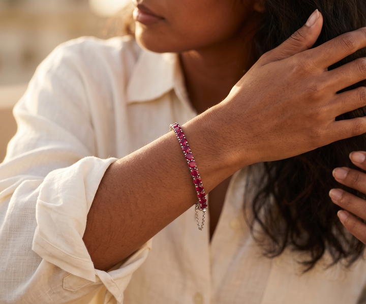 A close-up lifestyle shot of a woman wearing a delicate beaded bracelet with vibrant pinkish-red rubies and silver-toned chain links, paired with a white collared shirt.