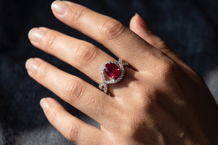 A close-up of a hand wearing a silver ring with a heart-shaped red gemstone surrounded by small clear stones.
