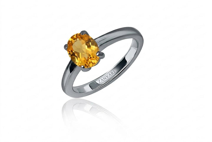 A studio product shot of a silver solitaire ring featuring a faceted oval yellow citrine in a four-prong setting, angled upright with a subtle mirror reflection on a clean white surface.