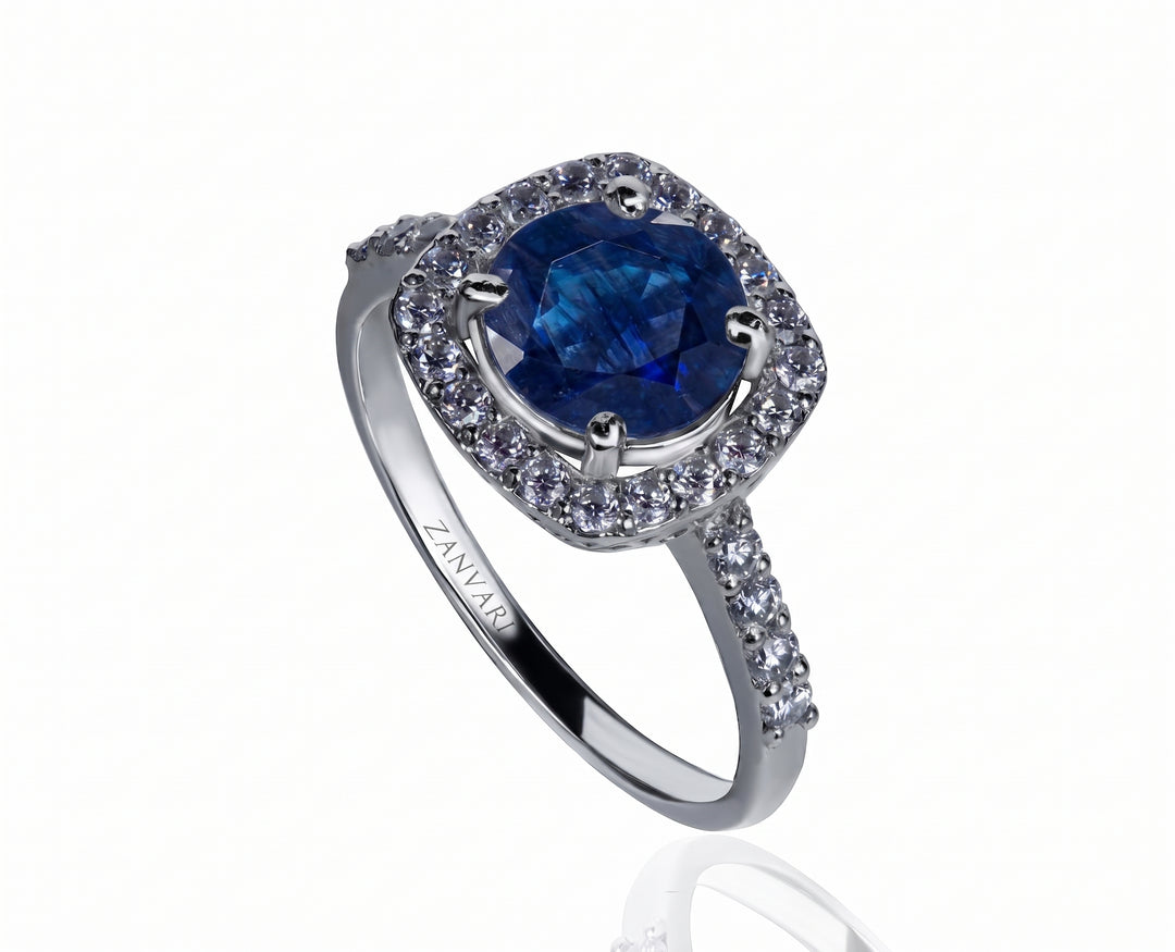 A silver-toned halo ring featuring a cushion-cut deep blue synthetic sapphire center stone and pavé-set clear stones along the shoulders, shown on a white background with a soft reflection.