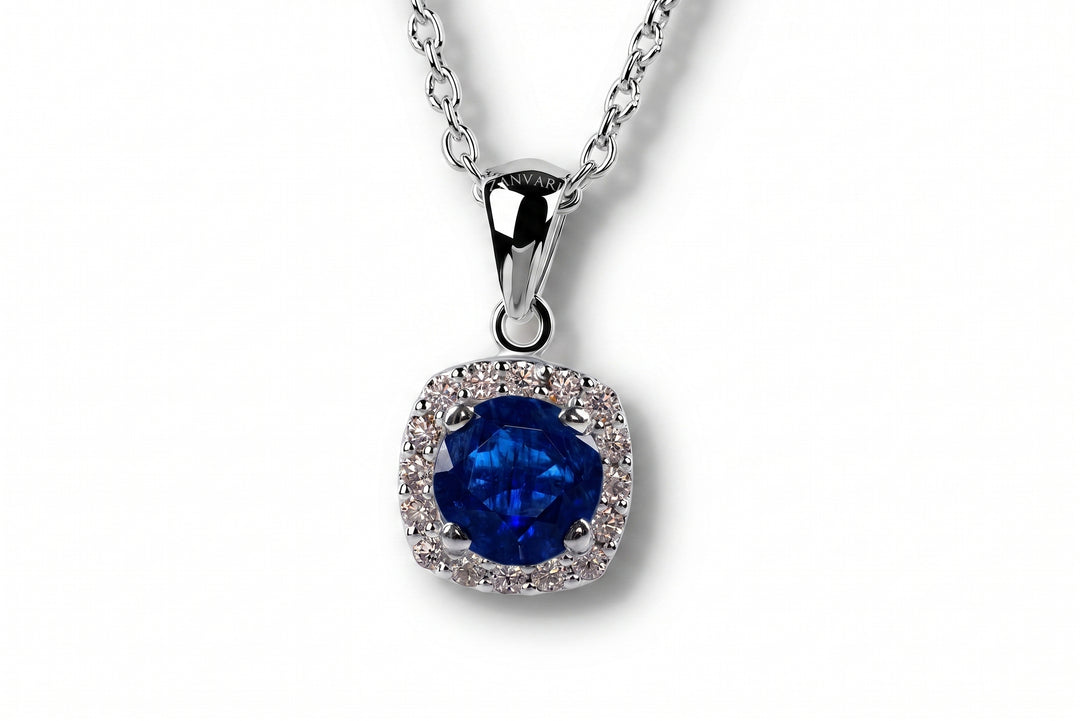 A silver pendant necklace featuring a cushion-cut deep blue synthetic sapphire gemstone surrounded by a halo of small clear stones, suspended from a silver chain on a white background.
