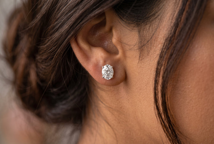Close-up macro shot of a woman's earlobe wearing a delicate oval-cut diamond solitaire stud earring in a white gold four-prong setting, with soft dark brown hair framing the side profile.