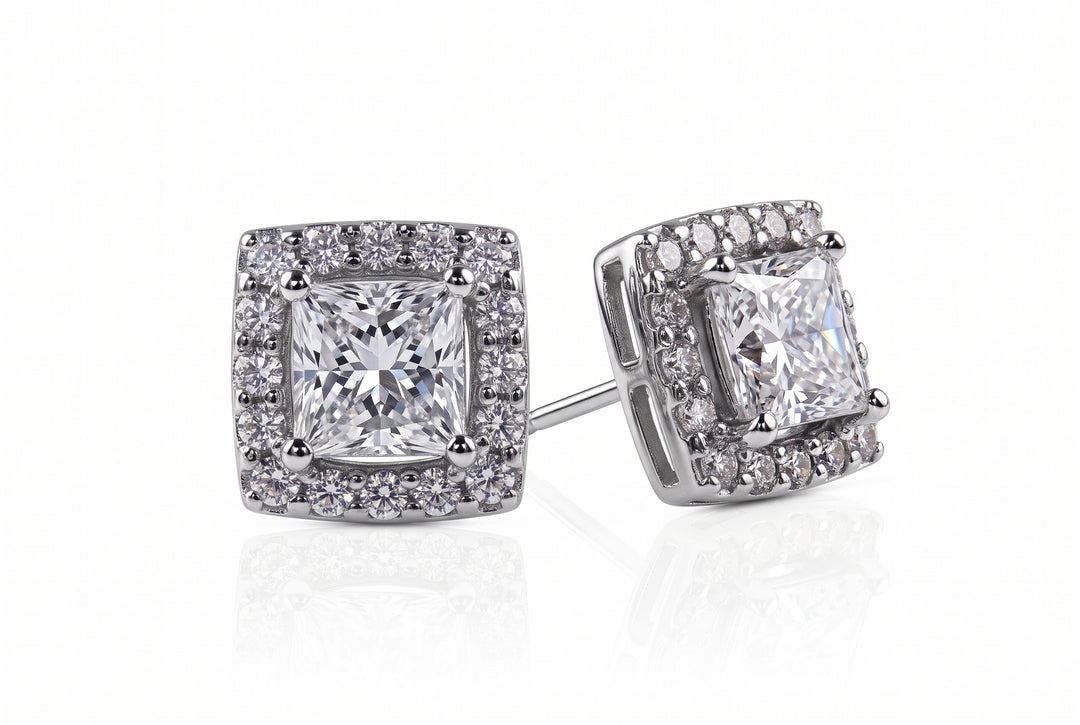 A pair of square halo stud earrings featuring a central princess-cut clear gemstone surrounded by a double halo of smaller round stones in a silver-toned metal setting, shown on a white background with a soft reflection.