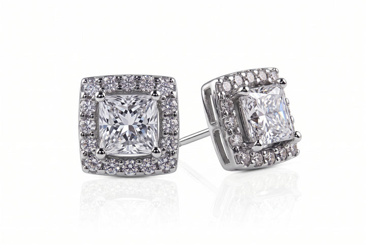 A pair of square halo stud earrings featuring a central princess-cut clear gemstone surrounded by a double halo of smaller round stones in a silver-toned metal setting, shown on a white background with a soft reflection.