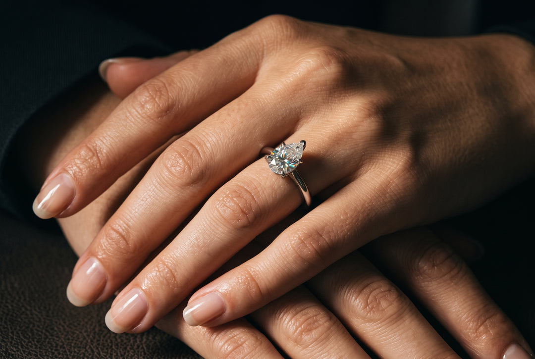 Close-up editorial photograph of a woman’s hand resting across another hand, wearing a refined, smaller-carat pear-cut moissanite solitaire ring on a slender white gold band under soft window lighting.