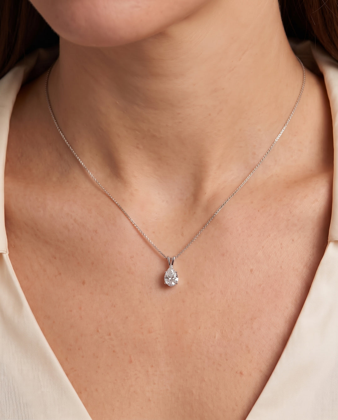 A close-up of a model wearing a delicate silver chain necklace with a pear-cut clear gemstone pendant, styled with a cream-colored top.