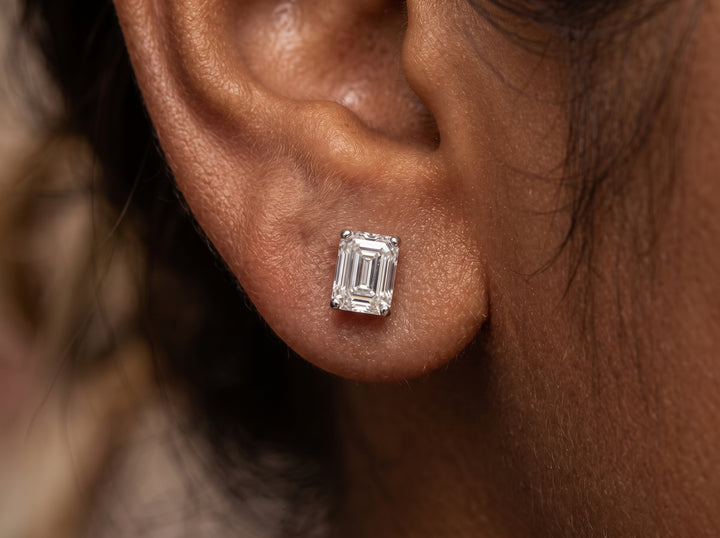 A macro photograph of a brown-skinned woman's earlobe featuring a small, rectangular emerald-cut diamond stud earring, with loose strands of dark hair visible around the ear.