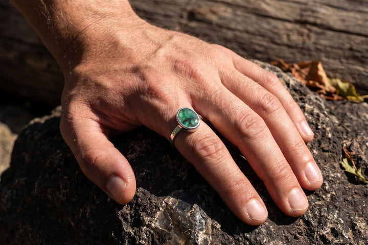 Close-up shot of a masculine male hand with defined knuckles and natural skin texture resting firmly on a dark, textured rock outdoors. The hand wears a polished silver bezel-set ring on the ring finger, featuring an oval, opaque natural green emerald with visible inclusions. Dramatic sunlight casts realistic highlights and shadows across the hand and setting, with a blurred natural wood background.
