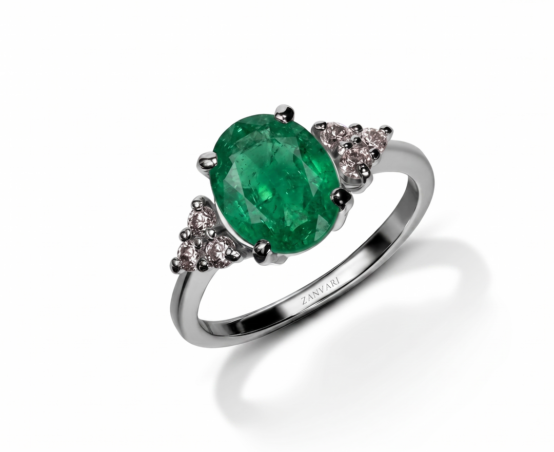 An oval-cut emerald green gemstone ring set in a polished silver-colored metal band, accented with small moissanite clear stones on each side. The brand name 'Zanvari' is engraved on the inner band.