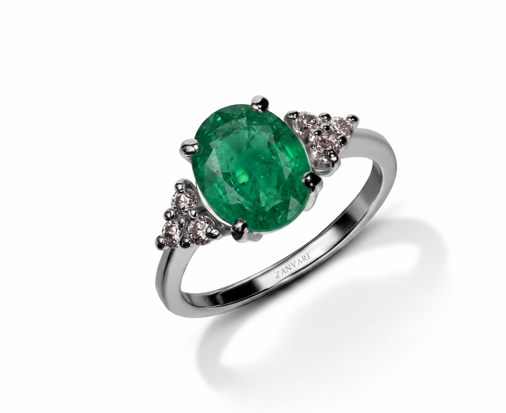 An oval-cut emerald green gemstone ring set in a polished silver-colored metal band, accented with small moissanite clear stones on each side. The brand name 'Zanvari' is engraved on the inner band.
