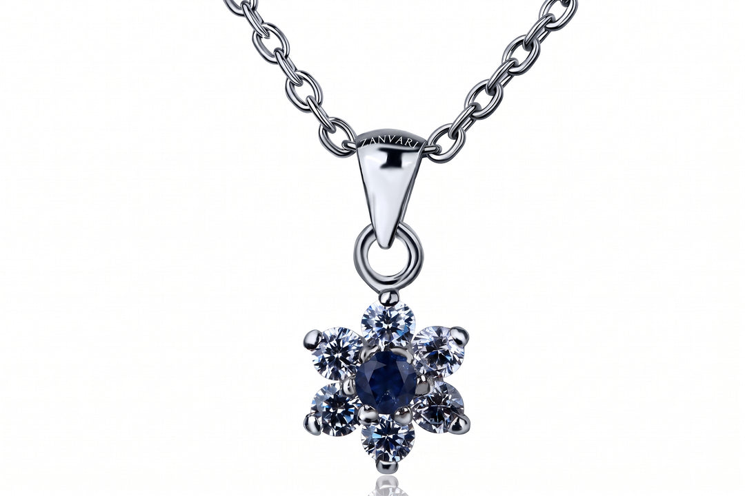 A silver-tone pendant necklace featuring a flower-shaped setting with a central dark blue oval stone surrounded by six clear round stones, suspended from a matching silver chain on a white background.