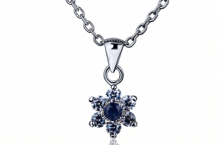 A silver-tone pendant necklace featuring a flower-shaped setting with a central dark blue oval stone surrounded by six clear round stones, suspended from a matching silver chain on a white background.