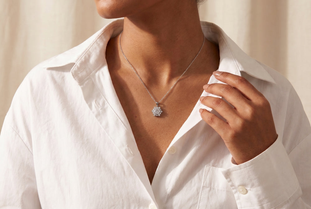 A close-up lifestyle shot of a model wearing a delicate silver chain necklace with a textured, flower-like circular pendant, styled with an open white button-down shirt against a warm beige background.