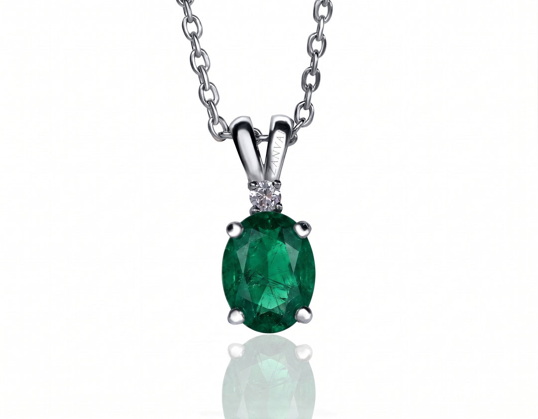 An oval-cut green emerald pendant necklace in a silver-toned setting with a small clear accent stone, suspended from a matching silver chain against a white background.
