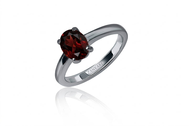 A studio product photograph of an elegant solitaire ring featuring a faceted oval-cut deep crimson-red garnet gemstone secured by four prominent prongs on a sleek, polished silver band with the "ZANVARI" engraving visible on the inside shank. The ring is angled upright against an isolated pure white background with a subtle mirror reflection beneath it.