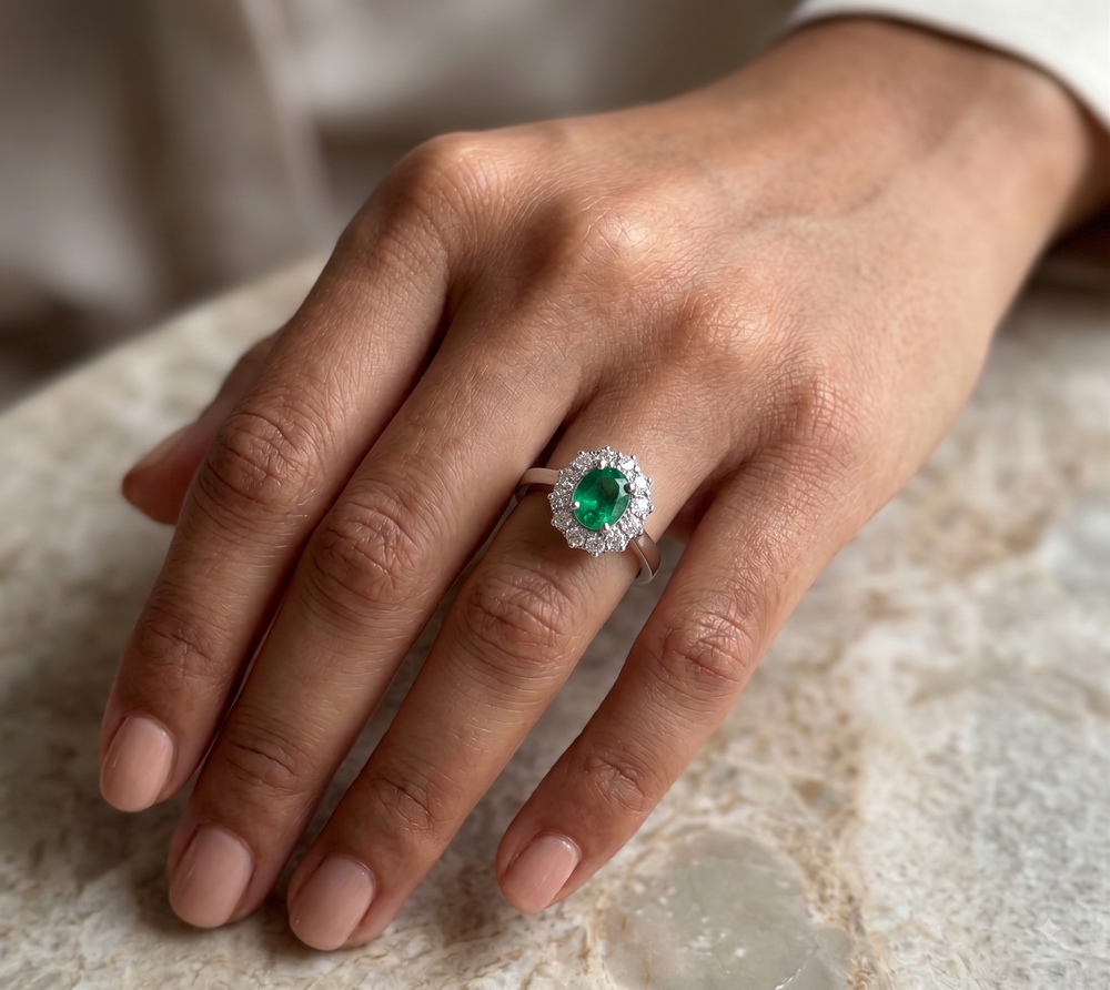 A close-up of a hand wearing a ring with a round green emerald-cut center stone surrounded by a halo of small clear stones on a warm-toned metal band, resting on a light stone surface.