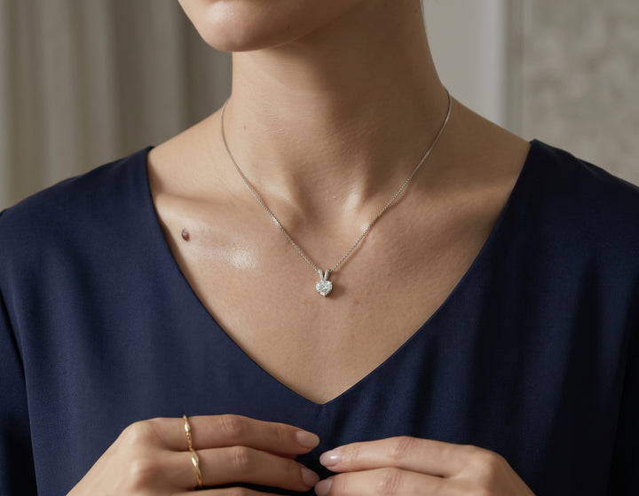 Close-up shot of a woman wearing a delicate silver chain necklace with a small, heart-shaped diamond pendant resting against her collarbone, set against a dark blue V-neck top.   
JPG